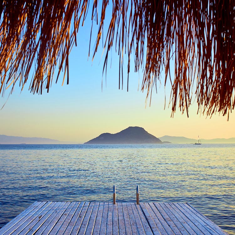 Der untere Teil eines Strohdaches (Sonnenschirm) hängt über dem oberen Bildrand. Im sanften Abendlicht erstreckt sich das Meer mit einer markanten, dunklen Insel in der Mitte und Bergen am Horizont.