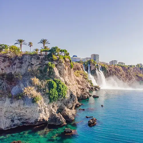 Eine Luftaufnahme des Duden-Wasserfalls, der von einer hohen Klippe ins türkisfarbene Mittelmeer in Antalya stürzt. Üppige Vegetation bedeckt die Klippen, während im Hintergrund Gebäude einer Stadt unter einem klaren blauen Himmel zu sehen sind.