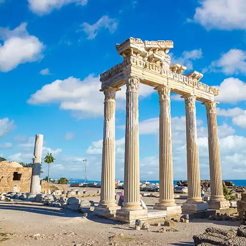 Fünf hohe, rekonstruierte korinthische Säulen des Apollon-Tempels in Side, Türkei, stehen vor dem klaren, blauen Mittelmeer. Am Boden befinden sich weitere Ruinen, unter einem strahlend blauen Himmel mit weißen Wolken.