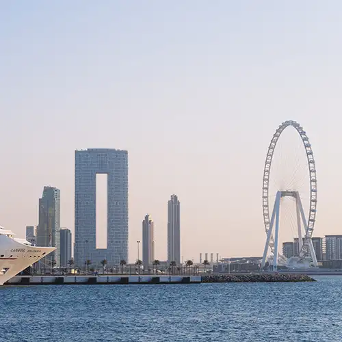 Dubai Marina Panorama mit Ain Dubai, modernen Wolkenkratzern und einem Kreuzfahrtschiff. Blick vom Meer auf die Skyline von Dubai. Rechts steht das Riesenrad Ain Dubai, links moderne Wolkenkratzer, darunter das Hochhaus mit dem mittigen Ausschnitt. Ein großes, weißes Kreuzfahrtschiff liegt am Kai.