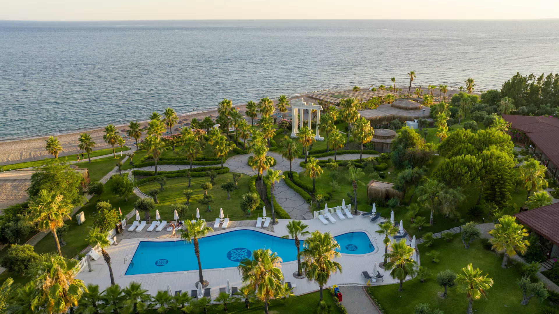 Poolanlage von Flora Garden Beach Hotel Hotel mit großen, menschenleeren Pool, an dem weiße Sonnenschirme stehen. Im Hintergrund ist Meer zu sehen.
