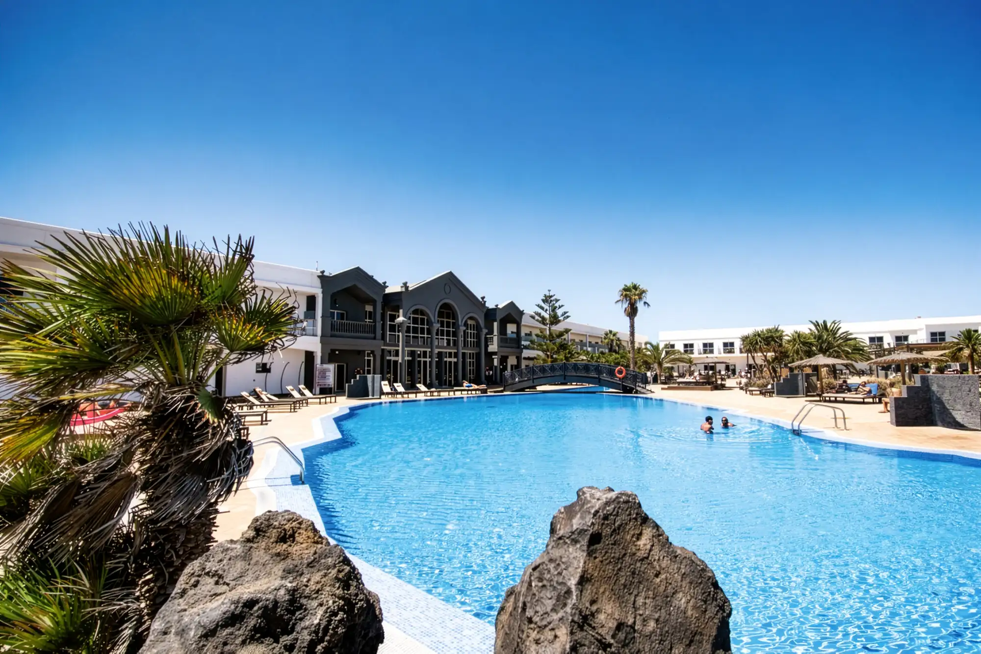 Coral Hotel Ein sonniger Blick über einen großen, geschwungenen Hotelpool auf einer Ferienanlage. Am Poolufer stehen Palmen und vulkanische Felsen. Im Hintergrund befinden sich die Hotelgebäude mit dunklerer, architektonisch interessanter Lobby, einer Brücke über den Pool und Liegestühlen. Ein Paar schwimmt im klaren blauen Wasser unter einem strahlend blauen Himmel.
